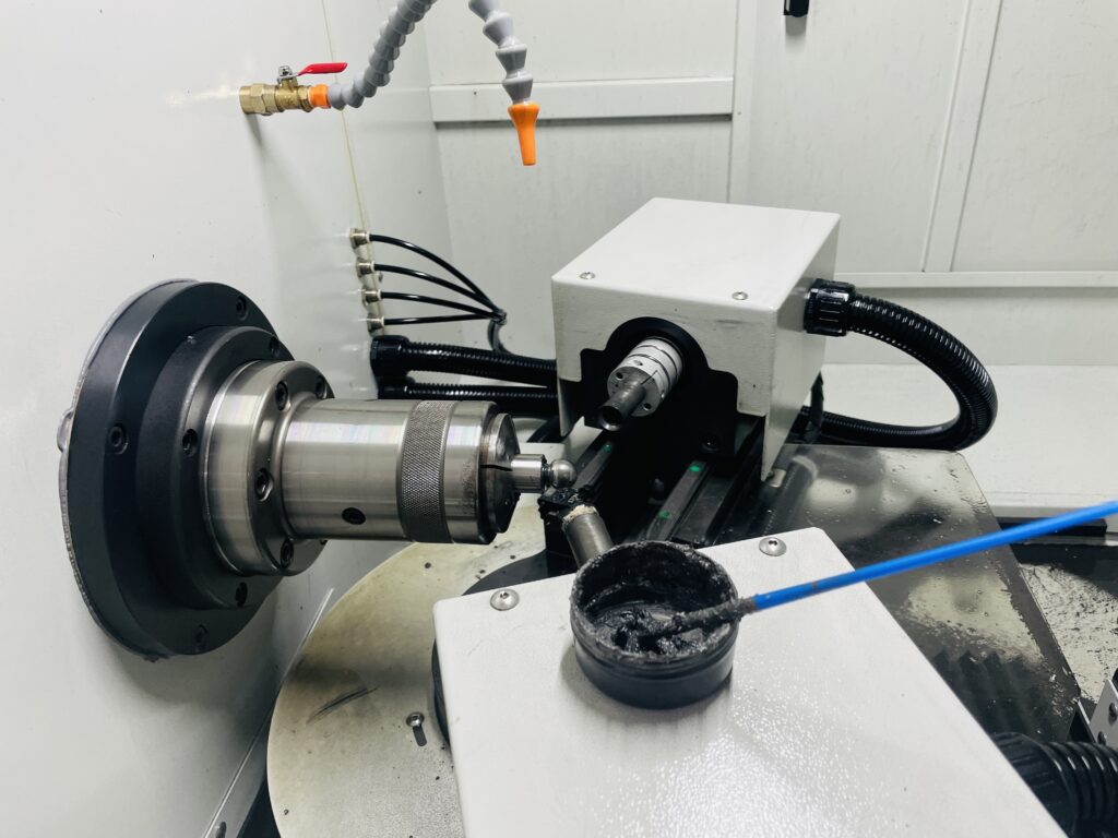Pump slipper ball-end grinding machine — Rohre Pump component ball end held in a grinding machine, with spindle, tooling and coolant nozzles visible.